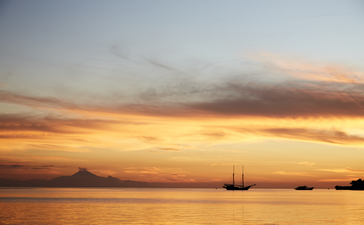 Sunset over calm waters at Amandira, with a sailing vessel silhouetted against golden and orange skies.