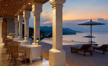 Amankila resort terrace at dusk, with dining area overlooking Bali's coastline and calm waters.