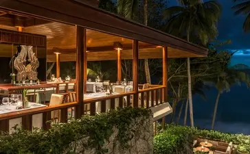 Amanpuri resort dining pavilion illuminated at dusk, overlooking tropical gardens and coastline.
