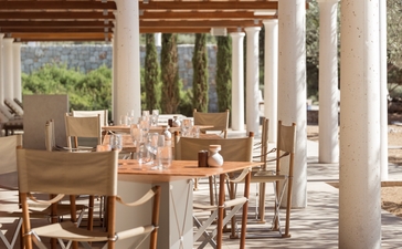 アマンゾイ・リゾートのビーチクラブ・ダイニングエリア。白い柱が並ぶテラスに木製テーブルと椅子が配置され、窓の先にはギリシャの海岸風景が広がる。
