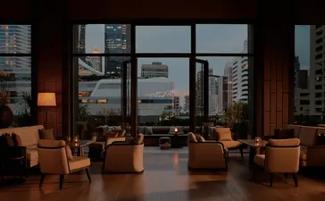 Terrace lounge at Aman Nai Lert Bangkok with floor-to-ceiling windows overlooking the city skyline at dusk.