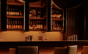 Aman Nai Lert Bangkok lounge bar with illuminated spirit bottles displayed behind polished wooden counter.