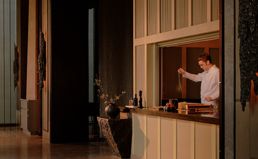 Two staff members work at the reception desk of Aman Nai Lert Bangkok, framed by tall wooden doors in a dimly lit corridor.