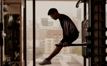 A man stretches in the gym at Aman Nai Lert Bangkok, silhouetted against floor-to-ceiling windows overlooking the city.