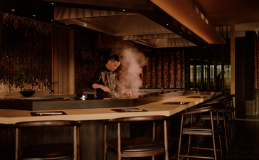 Warm-lit bar at Aman Nai Lert Bangkok with a server behind the counter and wooden interior details.