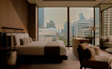 アマン ナイラート バンコクのホテル、ルーム145からバンコクの街並みを望む寝室。大きな窓から都市景観が見え、ベージュのベッドリネンと落ち着いた色調のインテリアが特徴。