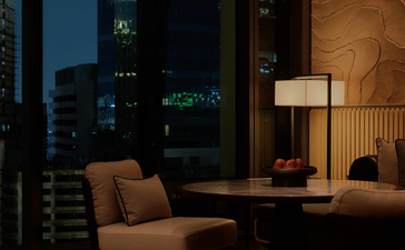 Mid-century modern armchair and side table in a Bangkok hotel room overlooking the city at dusk.