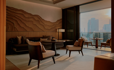 غرفة جلوس في أمان ناي ليرت بانكوك، تطل على أفق المدينة من خلال نوافذ كبيرة، مع كراسي جلوس وديكور دافئ.