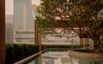 Swimming pool at Aman Nai Lert Bangkok with view of modern building and tree reflected in still water.