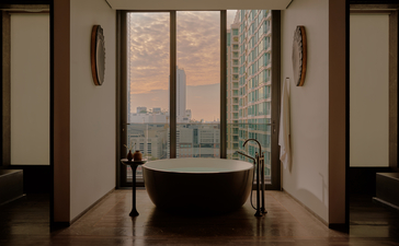 Aman Suite bathroom at Aman Nai Lert Bangkok, featuring a freestanding bathtub with city views through floor-to-ceiling windows.