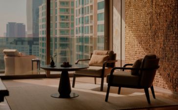 Aman Nai Lert Bangkok suite lounge with city views, featuring mid-century chairs and textured stone wall.