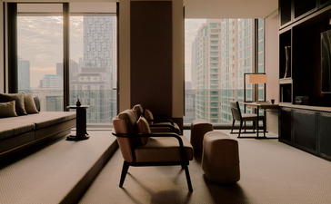 Aman Suite lounge at Aman Nai Lert Bangkok with floor-to-ceiling windows overlooking the city skyline, chocolate leather armchair, and minimalist furnishings.