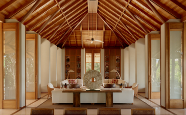 Wooden villa living area at Amanyara with vaulted ceiling, central dining table, and glass doors opening to natural light.