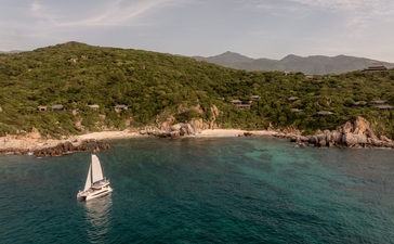 Aerial view of a catamaran sailing along the turquoise coast at Amanoi, Vietnam, with forested hillside beyond.