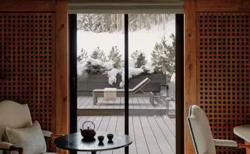 Bedroom at Aman Le Mélézin with view of snow-covered slopes through large window, terracotta-toned walls and modern furnishings.