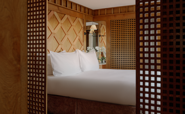 Chambre village bedroom at Aman Le Mélézin, viewed through wooden lattice screens, with white bedding and warm wood tones.