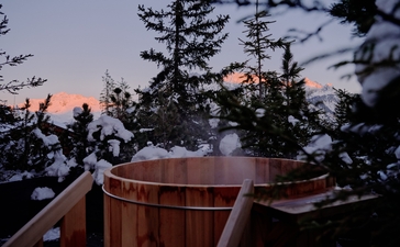 Wooden hot tub on a terrace at Aman Le Mélézin at dusk, with alpine forest and sky beyond.