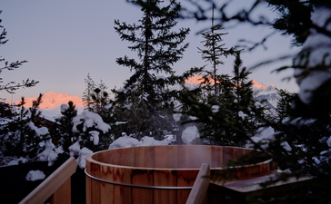 Wooden hot tub on a terrace at Aman Le Mélézin at dusk, with alpine forest and sky beyond.