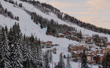 Skiers on snow-covered slopes at Aman Le Mélézin, with forested mountains and alpine buildings in the distance.