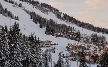 Skiers on snow-covered slopes at Aman Le Mélézin, with forested mountains and alpine buildings in the distance.
