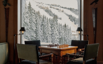 Salon at Aman Le Mélézin with wooden chairs at a desk facing a window overlooking snowy Alpine forest.