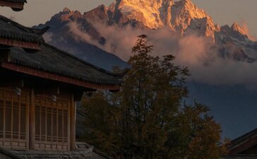 Golden sunrise light illuminates snow-capped mountain peaks above Amandayan's roofline, with mist rising through forested valleys below.