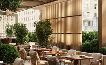 Terrazza del giardino di Aman New York con tavoli da pranzo in legno sotto una struttura coperta, circondata da piante e pareti in pietra.