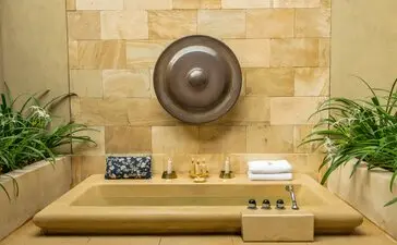 Menoreh Pavilion outdoor bath at Amanjiwo resort, featuring a sunken stone basin beneath a circular bronze fixture, flanked by potted plants and toiletries.
