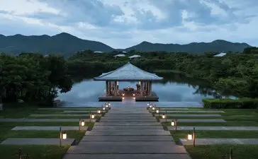 Wooden dining pavilion lit by lanterns on a lake at Amanoi, Vietnam, at dusk with forested mountains beyond.
