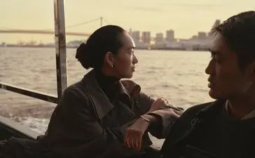 Two people conversing on a Sumida River cruise in Tokyo at dusk, with the city skyline visible across the water.