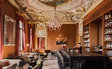 Aman Venice bar hall with ornate ceiling, red walls, crystal chandelier, and cocktail-making counter.