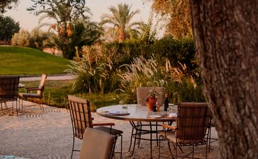Breakfast terrace beneath olive trees at Amanjena, Morocco, with wooden chairs and tables on a stone patio.