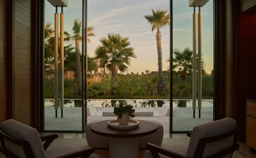 Amanvari Estuary Casita living area with seated figures overlooking palm trees and waterway through open glass doors.