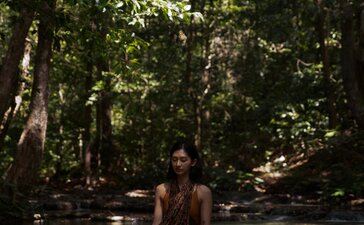 Woman meditating in river at Amanwana, Indonesia, surrounded by forest canopy.