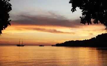 Sunset over Amanwana's coastline, with sailing boats silhouetted against golden and amber skies reflecting on calm waters.