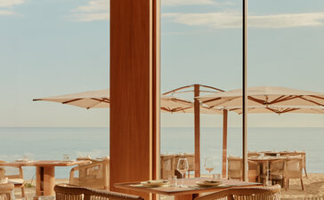 Amanvari interior lounge with wooden furniture and expansive city views through floor-to-ceiling windows.