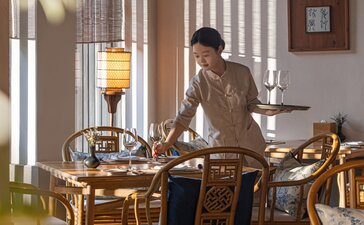 Resident at Amanfayun seated at wooden desk beside tall windows with vertical blinds, warm natural light streaming through.