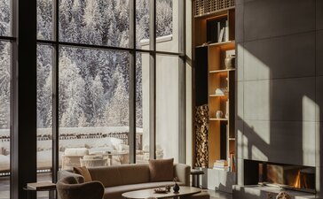 Sitting area at Aman Rosa Alpina resort with floor-to-ceiling windows framing alpine views, two leather chairs, and warm wooden accents.