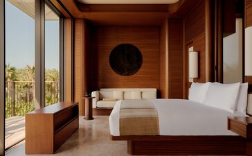 Amanvari casita bedroom with wooden furnishings, large windows overlooking desert landscape, and minimal décor.