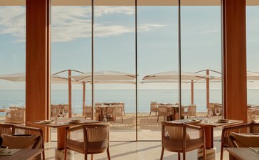 Amanvari dining venue with wooden beams, chairs, and expansive windows overlooking desert landscape.