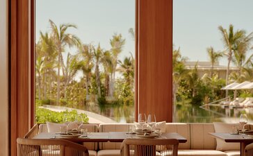 Amanvari dining area with wooden table and chairs overlooking palm-lined gardens through floor-to-ceiling windows.