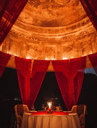 Candlelit table for two beneath ornate golden dome at Amanbagh, framed by red curtains.