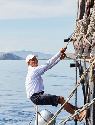 Crew member scaling the rigging of Amandira yacht with calm waters and distant islands beyond.