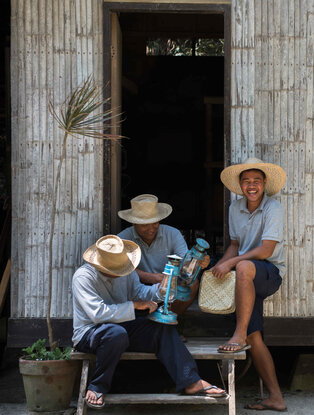 Two men sit on a wooden bench outside a weathered wooden doorway at Amanpulo, one wearing a straw hat and light shirt, the other in blue clothing.