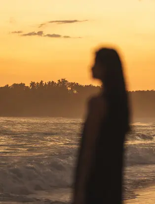 Silhouetted figure standing on Amanera's beach at sunset, golden sky reflected in calm water.