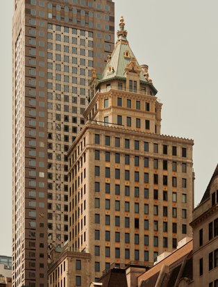 Fachada de edifício histórico com detalhes arquitetônicos dourados ao lado de torre moderna em Aman New York.