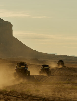 Three off-road vehicles traverse dusty desert terrain at Amangiri resort, with a striking rock formation rising in the background.