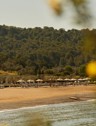 Amanzoe beach club with golden sand, tree-lined shore and calm waters beneath cloudy sky.