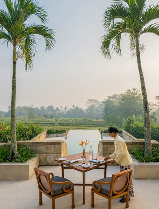 Two wicker chairs at a table on a terrace at Amanjiwo, overlooking rice fields and distant mountains in Bali, Indonesia.