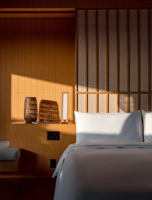 Amanemu bedroom with wooden wall panels, white bed, and warm natural light filtering through vertical screens.